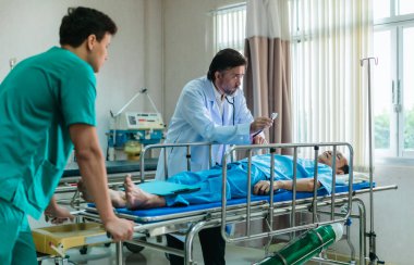 Kıdemli hekim hastane yatağında yatan hastayı muayene etmek için steteskop kullanırken, bir doktor dikkatlice gözlemliyor. Kurulum, profesyonel tıp ortamında acil durum ve takım çalışmasını iletiyor