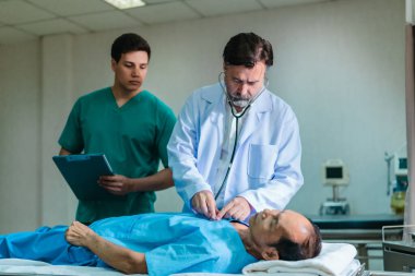 Kıdemli hekim hastane yatağında yatan hastayı muayene etmek için steteskop kullanırken, bir doktor dikkatlice gözlemliyor. Kurulum, profesyonel tıp ortamında acil durum ve takım çalışmasını iletiyor