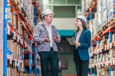 Kadın kalite kontrol müdürü, dağıtım merkezindeki erkek depo yöneticisiyle konuşurken envanter kontrolü yapıyor. Takım çalışması, kalite güvencesi, profesyonel depo operasyonları.