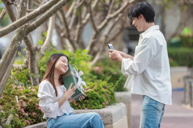 Asyalı bir çift, açık havada hediye kutularını değiş tokuş ederken neşeli bir anı paylaşıyor. Sahne, özel bir durum sırasında sevgi, kutlama ve bağlantı duygularını yakalar..