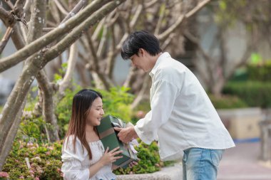 Asyalı bir çift, açık havada hediye kutularını değiş tokuş ederken neşeli bir anı paylaşıyor. Sahne, özel bir durum sırasında sevgi, kutlama ve bağlantı duygularını yakalar..