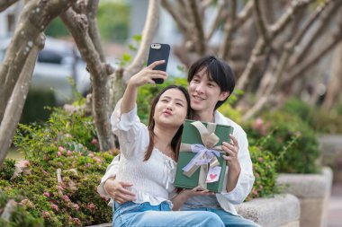 Asyalı bir çift, açık havada hediye kutularını değiş tokuş ederken neşeli bir anı paylaşıyor. Sahne, özel bir durum sırasında sevgi, kutlama ve bağlantı duygularını yakalar..