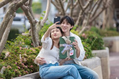 Asyalı bir çift, açık havada hediye kutularını değiş tokuş ederken neşeli bir anı paylaşıyor. Sahne, özel bir durum sırasında sevgi, kutlama ve bağlantı duygularını yakalar..