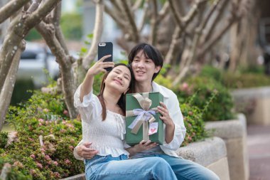 Asyalı bir çift, açık havada hediye kutularını değiş tokuş ederken neşeli bir anı paylaşıyor. Sahne, özel bir durum sırasında sevgi, kutlama ve bağlantı duygularını yakalar..