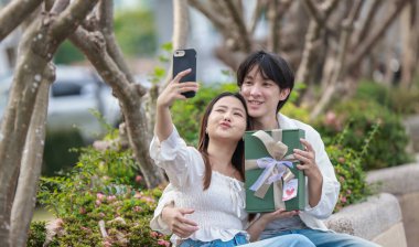 Asyalı bir çift, açık havada hediye kutularını değiş tokuş ederken neşeli bir anı paylaşıyor. Sahne, özel bir durum sırasında sevgi, kutlama ve bağlantı duygularını yakalar..