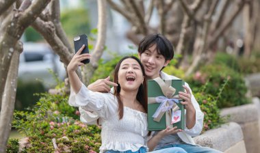 Asyalı bir çift, açık havada hediye kutularını değiş tokuş ederken neşeli bir anı paylaşıyor. Sahne, özel bir durum sırasında sevgi, kutlama ve bağlantı duygularını yakalar..