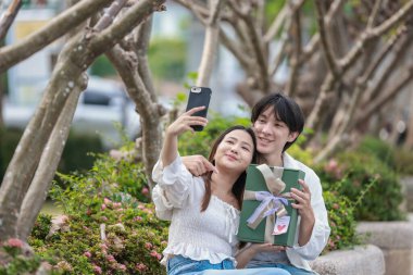 Asyalı bir çift, açık havada hediye kutularını değiş tokuş ederken neşeli bir anı paylaşıyor. Sahne, özel bir durum sırasında sevgi, kutlama ve bağlantı duygularını yakalar..