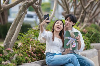 Asyalı bir çift, açık havada hediye kutularını değiş tokuş ederken neşeli bir anı paylaşıyor. Sahne, özel bir durum sırasında sevgi, kutlama ve bağlantı duygularını yakalar..