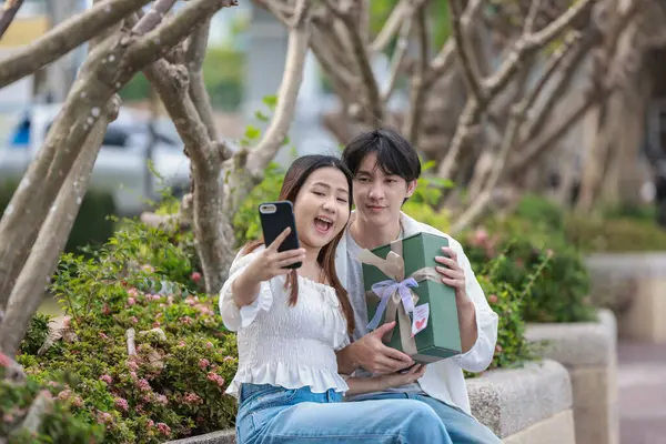 Asyalı bir çift, açık havada hediye kutularını değiş tokuş ederken neşeli bir anı paylaşıyor. Sahne, özel bir durum sırasında sevgi, kutlama ve bağlantı duygularını yakalar..