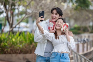 Asyalı bir çift, açık havada hediye kutularını değiş tokuş ederken neşeli bir anı paylaşıyor. Sahne, özel bir durum sırasında sevgi, kutlama ve bağlantı duygularını yakalar..