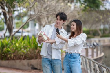 Asyalı bir çift, açık havada hediye kutularını değiş tokuş ederken neşeli bir anı paylaşıyor. Sahne, özel bir durum sırasında sevgi, kutlama ve bağlantı duygularını yakalar..