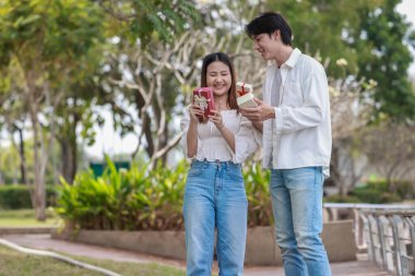 Asyalı bir çift, açık havada hediye kutularını değiş tokuş ederken neşeli bir anı paylaşıyor. Sahne, özel bir durum sırasında sevgi, kutlama ve bağlantı duygularını yakalar..