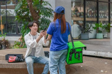 Asyalı adam akıllı telefonu kadın kuryeden teslimat alırken nakitsiz ödeme yapmak için kullanıyor. Modern işlemler açık havada gerçekleşir, ulaşılamayan hizmetlerin kolaylığı ve verimliliği yakalanır.