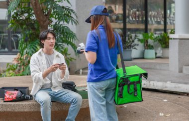 Asyalı adam akıllı telefonu kadın kuryeden teslimat alırken nakitsiz ödeme yapmak için kullanıyor. Modern işlemler açık havada gerçekleşir, ulaşılamayan hizmetlerin kolaylığı ve verimliliği yakalanır.