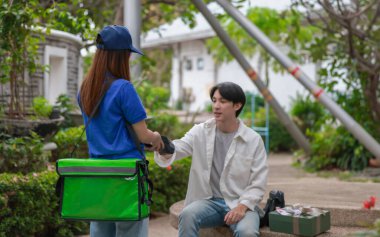 Asyalı adam akıllı telefonu kadın kuryeden teslimat alırken nakitsiz ödeme yapmak için kullanıyor. Modern işlemler açık havada gerçekleşir, ulaşılamayan hizmetlerin kolaylığı ve verimliliği yakalanır.