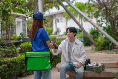 Asyalı adam akıllı telefonu kadın kuryeden teslimat alırken nakitsiz ödeme yapmak için kullanıyor. Modern işlemler açık havada gerçekleşir, ulaşılamayan hizmetlerin kolaylığı ve verimliliği yakalanır.