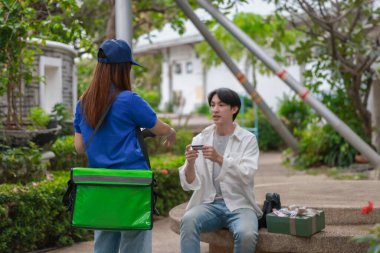 Asyalı adam akıllı telefonu kadın kuryeden teslimat alırken nakitsiz ödeme yapmak için kullanıyor. Modern işlemler açık havada gerçekleşir, ulaşılamayan hizmetlerin kolaylığı ve verimliliği yakalanır.