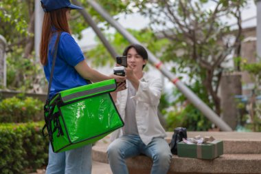 Asyalı adam akıllı telefonu kadın kuryeden teslimat alırken nakitsiz ödeme yapmak için kullanıyor. Modern işlemler açık havada gerçekleşir, ulaşılamayan hizmetlerin kolaylığı ve verimliliği yakalanır.