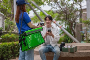 Asyalı adam akıllı telefonu kadın kuryeden teslimat alırken nakitsiz ödeme yapmak için kullanıyor. Modern işlemler açık havada gerçekleşir, ulaşılamayan hizmetlerin kolaylığı ve verimliliği yakalanır.