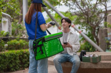 Asyalı adam akıllı telefonu kadın kuryeden teslimat alırken nakitsiz ödeme yapmak için kullanıyor. Modern işlemler açık havada gerçekleşir, ulaşılamayan hizmetlerin kolaylığı ve verimliliği yakalanır.