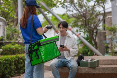 Asyalı adam akıllı telefonu kadın kuryeden teslimat alırken nakitsiz ödeme yapmak için kullanıyor. Modern işlemler açık havada gerçekleşir, ulaşılamayan hizmetlerin kolaylığı ve verimliliği yakalanır.