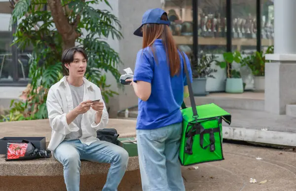 Asyalı adam akıllı telefonu kadın kuryeden teslimat alırken nakitsiz ödeme yapmak için kullanıyor. Modern işlemler açık havada gerçekleşir, ulaşılamayan hizmetlerin kolaylığı ve verimliliği yakalanır.
