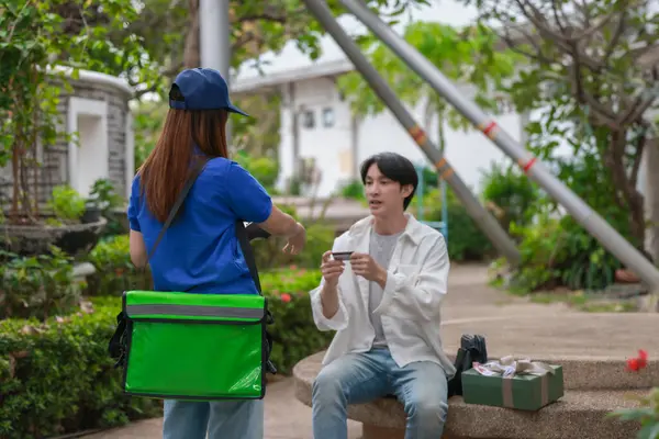 Asyalı adam akıllı telefonu kadın kuryeden teslimat alırken nakitsiz ödeme yapmak için kullanıyor. Modern işlemler açık havada gerçekleşir, ulaşılamayan hizmetlerin kolaylığı ve verimliliği yakalanır.