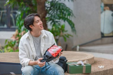 Asyalı bir adam elinde bir sürü hediye kutusu ve bir buket kırmızı gül tutarak sevinç ve heyecan ifade ederken sıcak bir şekilde gülümser. Romantik ve düşünceli bir jest yakalamak, kutlama için mükemmel..