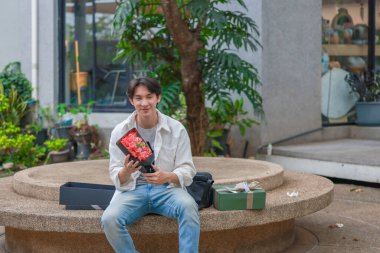 Asyalı bir adam elinde bir sürü hediye kutusu ve bir buket kırmızı gül tutarak sevinç ve heyecan ifade ederken sıcak bir şekilde gülümser. Romantik ve düşünceli bir jest yakalamak, kutlama için mükemmel..