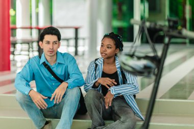 Farklı kültürlerden iki üniversite öğrencisi kampüs basamaklarında oturuyor ve elektrikli scooterın yanında arkadaşça sohbet ediyorlar. Sürdürülebilir şehir seyahati ve modern öğrenci hayatı