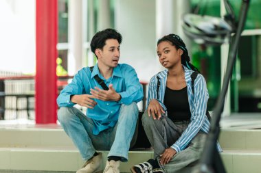 Farklı kültürlerden iki üniversite öğrencisi kampüs basamaklarında oturuyor ve elektrikli scooterın yanında arkadaşça sohbet ediyorlar. Sürdürülebilir şehir seyahati ve modern öğrenci hayatı