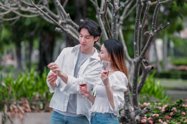 Asyalı çift kırmızı şarap ve sohbetten zevk alarak neşeli ve samimi bir anı paylaşıyor. Çiçekler ve yeşilliklerle çevrili. Romantizm, rahatlama ve bağlantı yakalamak