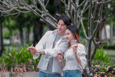 Asyalı çift kırmızı şarap ve sohbetten zevk alarak neşeli ve samimi bir anı paylaşıyor. Çiçekler ve yeşilliklerle çevrili. Romantizm, rahatlama ve bağlantı yakalamak