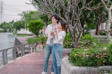 Asyalı çift kırmızı şarap ve sohbetten zevk alarak neşeli ve samimi bir anı paylaşıyor. Çiçekler ve yeşilliklerle çevrili. Romantizm, rahatlama ve bağlantı yakalamak