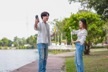 Neşeli bir Asyalı çift güneşli bir günde göl kenarında baloncuk çıkarmayı sever. Neşeli an, onların neşeli etkileşimlerini ve kaygısız ilişkilerini doğal bir ortamda yakalar..