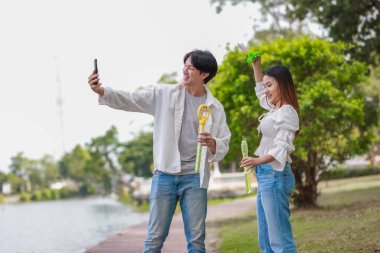 Neşeli bir Asyalı çift güneşli bir günde göl kenarında baloncuk çıkarmayı sever. Neşeli an, onların neşeli etkileşimlerini ve kaygısız ilişkilerini doğal bir ortamda yakalar..