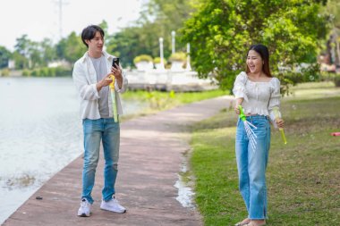 Neşeli bir Asyalı çift güneşli bir günde göl kenarında baloncuk çıkarmayı sever. Neşeli an, onların neşeli etkileşimlerini ve kaygısız ilişkilerini doğal bir ortamda yakalar..