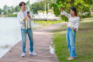 Neşeli bir Asyalı çift güneşli bir günde göl kenarında baloncuk çıkarmayı sever. Neşeli an, onların neşeli etkileşimlerini ve kaygısız ilişkilerini doğal bir ortamda yakalar..