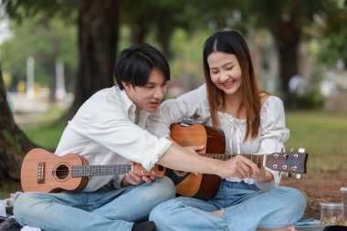 Asyalı çift piknik sırasında parkta dinlenmeyi, gülümsemeyi gitar çalmayı ve ukulele çalmayı seviyor. Doğal ortamda neşeli ve samimi bir bağlantı, müzik ve eğlence anı yakalıyorum..