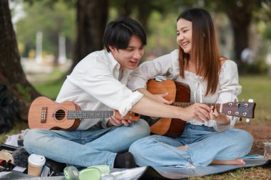 Asyalı çift piknik sırasında parkta dinlenmeyi, gülümsemeyi gitar çalmayı ve ukulele çalmayı seviyor. Doğal ortamda neşeli ve samimi bir bağlantı, müzik ve eğlence anı yakalıyorum..
