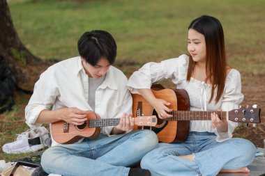 Asyalı çift piknik sırasında parkta dinlenmeyi, gülümsemeyi gitar çalmayı ve ukulele çalmayı seviyor. Doğal ortamda neşeli ve samimi bir bağlantı, müzik ve eğlence anı yakalıyorum..