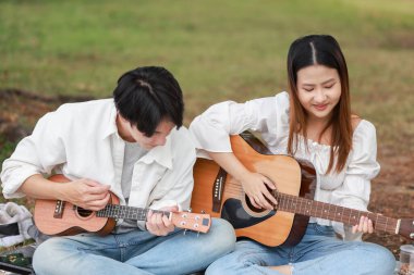 Asyalı çift piknik sırasında parkta dinlenmeyi, gülümsemeyi gitar çalmayı ve ukulele çalmayı seviyor. Doğal ortamda neşeli ve samimi bir bağlantı, müzik ve eğlence anı yakalıyorum..