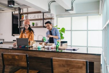 İki girişimci, modern bir çalışma alanında sürdürülebilir ambalaj örneklerini gözden geçiriyorlar. Çevre dostu materyaller ve ürün prototipleriyle çevrelenmiş olarak ürün geliştirme ve markalaşma fikirlerini tartışıyorlar.