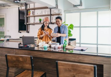 İki girişimci, modern bir çalışma alanında sürdürülebilir ambalaj örneklerini gözden geçiriyorlar. Çevre dostu materyaller ve ürün prototipleriyle çevrelenmiş olarak ürün geliştirme ve markalaşma fikirlerini tartışıyorlar.