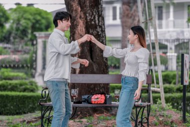 Asyalı çift, parktaki bankta eşine bir buket kırmızı çiçek ve hediye paketi sunan adamla samimi bir anın tadını çıkarıyor. Sıcak, açık hava manzarası aşkı, takdiri ve romantik kutlamayı yansıtıyor.