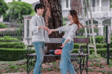Asyalı çift, parktaki bankta eşine bir buket kırmızı çiçek ve hediye paketi sunan adamla samimi bir anın tadını çıkarıyor. Sıcak, açık hava manzarası aşkı, takdiri ve romantik kutlamayı yansıtıyor.