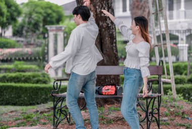 Asyalı çift, parktaki bankta eşine bir buket kırmızı çiçek ve hediye paketi sunan adamla samimi bir anın tadını çıkarıyor. Sıcak, açık hava manzarası aşkı, takdiri ve romantik kutlamayı yansıtıyor.