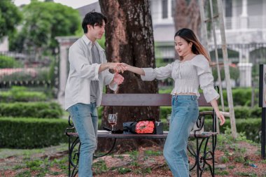Asyalı çift, parktaki bankta eşine bir buket kırmızı çiçek ve hediye paketi sunan adamla samimi bir anın tadını çıkarıyor. Sıcak, açık hava manzarası aşkı, takdiri ve romantik kutlamayı yansıtıyor.