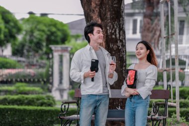 Asyalı çift, parktaki bankta eşine bir buket kırmızı çiçek ve hediye paketi sunan adamla samimi bir anın tadını çıkarıyor. Sıcak, açık hava manzarası aşkı, takdiri ve romantik kutlamayı yansıtıyor.