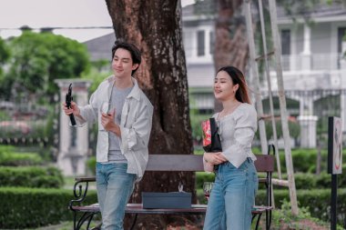 Asyalı çift, parktaki bankta eşine bir buket kırmızı çiçek ve hediye paketi sunan adamla samimi bir anın tadını çıkarıyor. Sıcak, açık hava manzarası aşkı, takdiri ve romantik kutlamayı yansıtıyor.